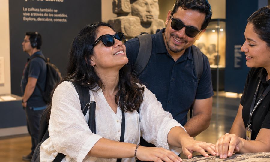 Una mujer con lentes oscuros y bastón blanco sonríe mientras recorre con las manos un relieve de piedra en un museo. Un hombre con lentes oscuros la acompaña y una guía del museo toca la piedra a su lado. Al fondo hay una escultura de piedra con rostro humano y un letrero que dice “Un legado que se siente”.